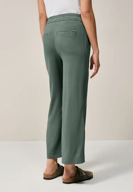 Frau trägt grüne Hose mit Gummizug und Sandalen, Ansicht von hinten.